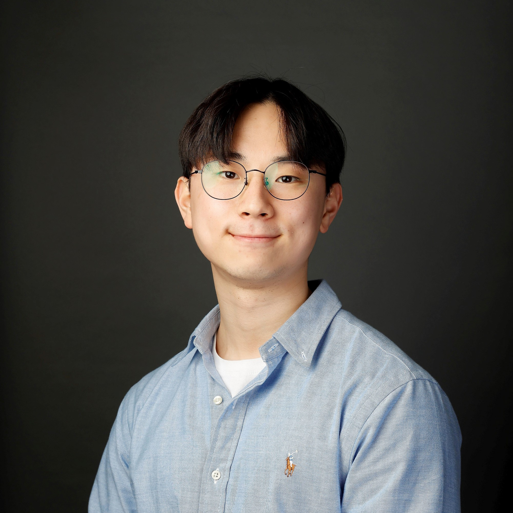 Portrait of a young man wearing round glasses and a light blue shirt against a dark background, facing the camera.