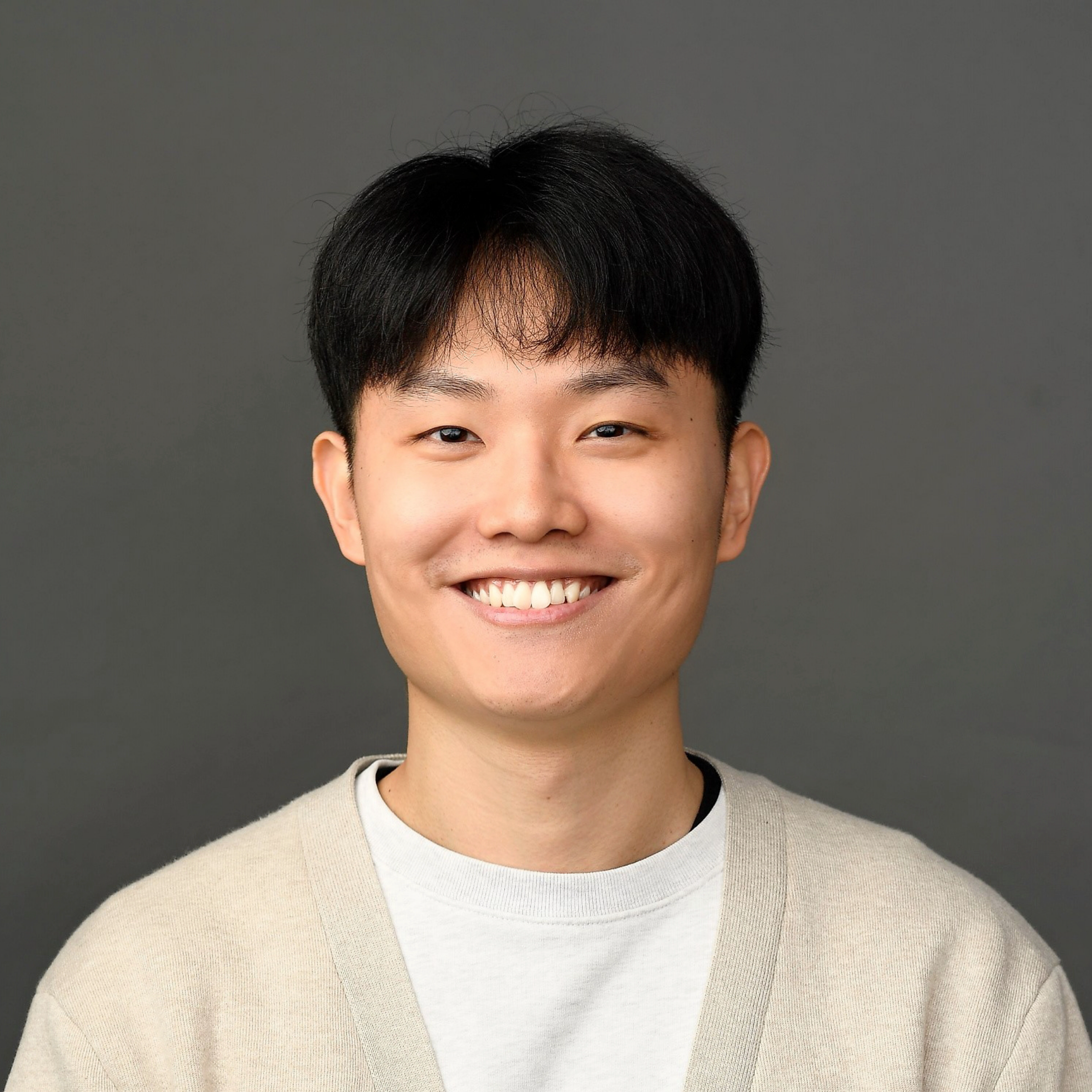 Young man smiling, only the lower half of his face visible, wearing a beige cardigan over a white shirt against a gray backdrop