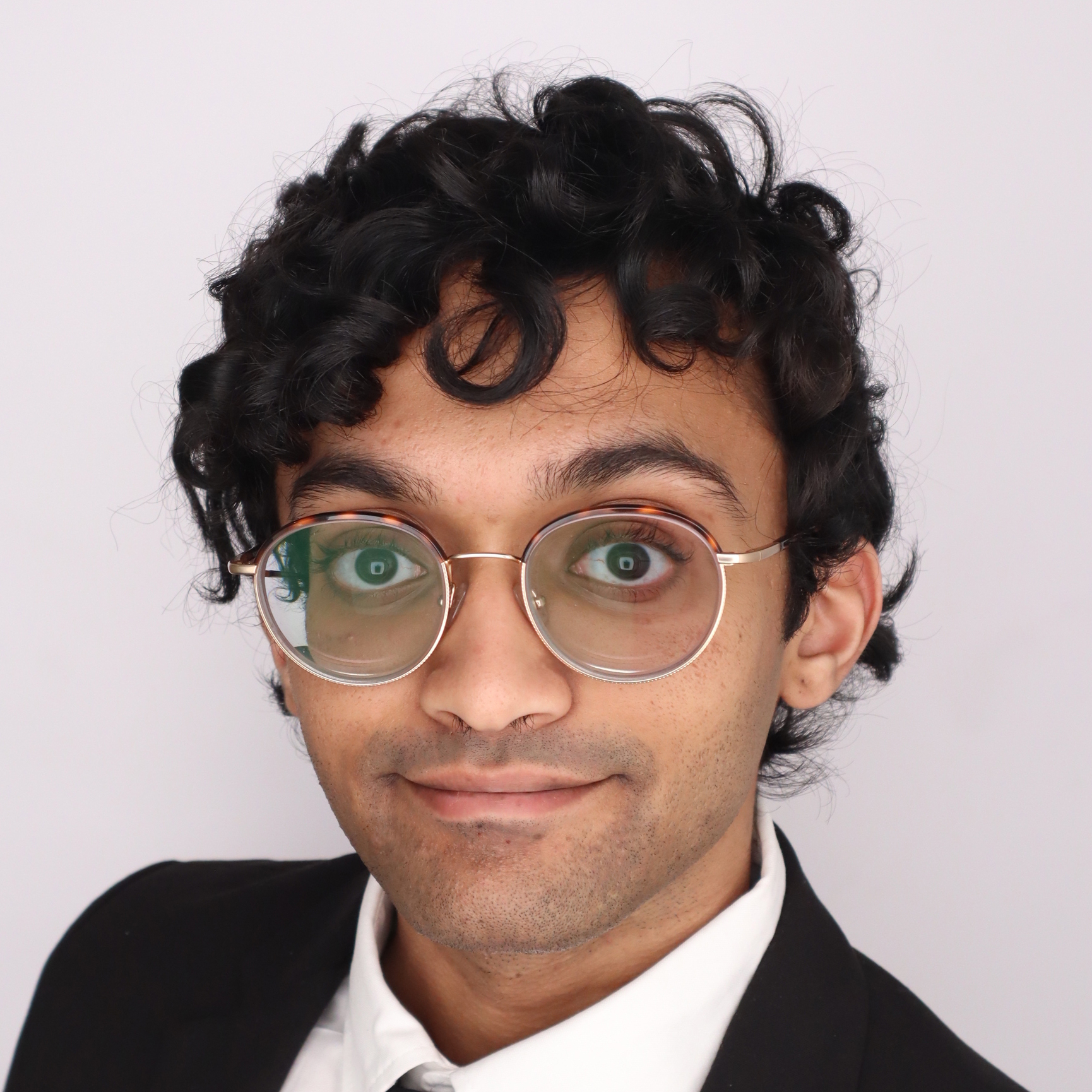 Close-up portrait of a person with curly black hair, round metal glasses, wearing a black blazer and white shirt, smiling subtly against a plain background.